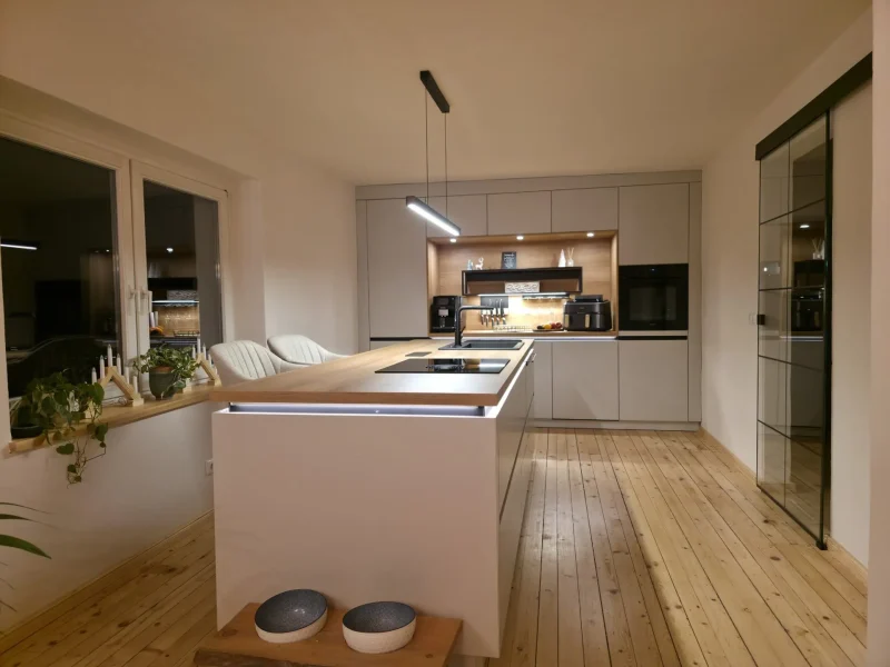 Moderne, zeitlos schöne Küche mit Holzfußboden, großer weißer Insel mit Barhockern, eingebautem Backofen und Kaffeemaschine, Unterschrankbeleuchtung, Topfpflanzen auf der Fensterbank und einem Glasschrank auf der rechten Seite.