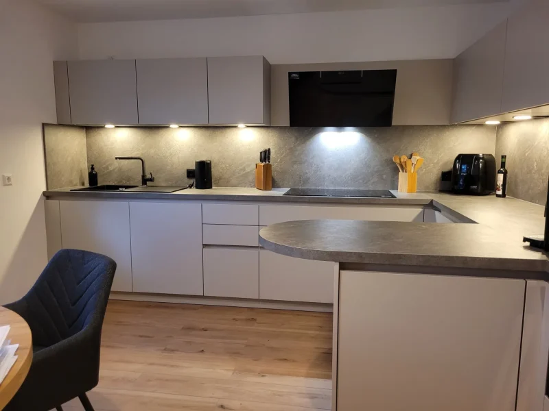 Moderne, wandelbare Küche mit grauen Schränken, Unterschrankbeleuchtung, schwarzer Spüle, Elektroherd, Messern, Geschirr, Kaffeemaschine und hellem Holzfußboden - ein zeitlos schöner Raum. Im Vordergrund sind ein dunkler Stuhl und ein runder Tisch zu sehen.