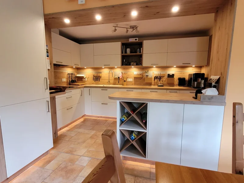 Moderne, wandelbare Küche mit weißen Schränken, Holzakzenten, Unterschrankbeleuchtung und Einbaugeräten. Auf der Insel befindet sich ein offenes Weinregal. Verschiedene Utensilien, eine Kaffeemaschine und Küchengeräte sind in dieser zeitlos schönen Umgebung übersichtlich angeordnet.