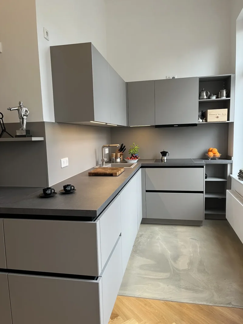 Moderne Küche mit eleganten grauen Schränken, schwarzen Arbeitsplatten und Einbaugeräten. Diese helle, helle Küche ist zeitlos schön, hell und luftig, mit minimalistischem Dekor - Pflanzen und Orangen verleihen dem sauberen, einladenden Raum einen frischen Touch.