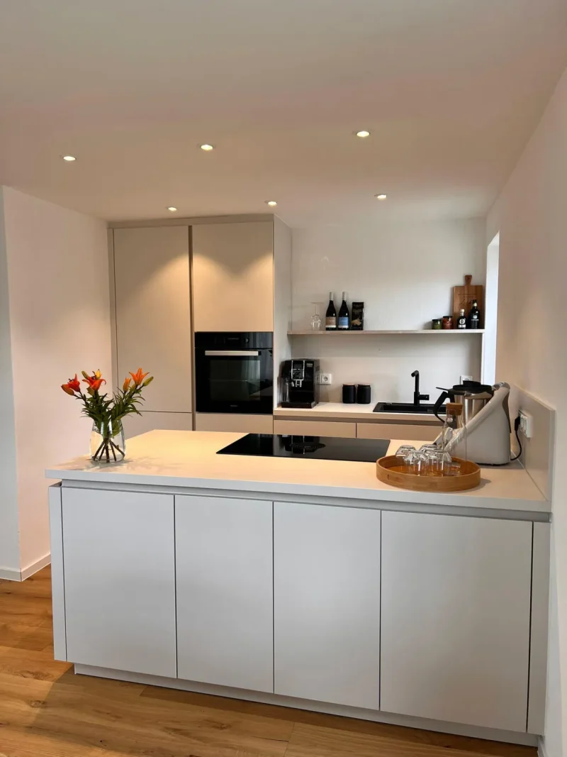 Moderne, zeitlos schöne Küche mit weißen Schränken, Einbaubackofen, Induktionskochfeld, offenen Regalen mit Gläsern und Flaschen, einer Vase mit Orangenblüten und einem Tablett mit Gläsern auf einem hellen Holzboden. Helle Küchen in minimalistischem Design.