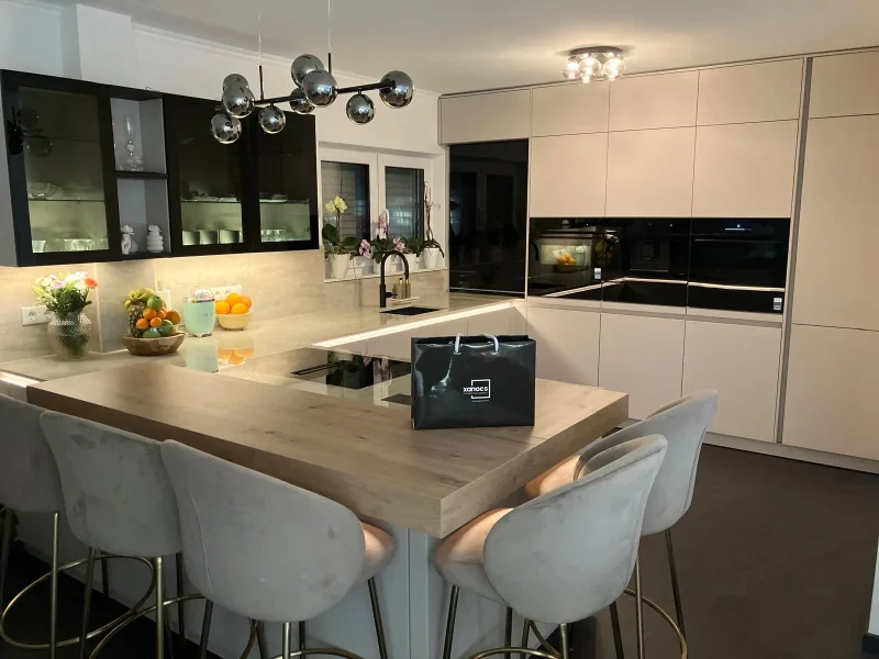 Moderne, zeitlos schöne Küche mit beigen Schränken, schwarzen Geräten, einer Holzinsel mit vier grauen Samtbarhockern und einer schwarzen Einkaufstasche auf der Insel. Obstschalen und dekorative Leuchten bereichern dieses helle Küchen-Design.