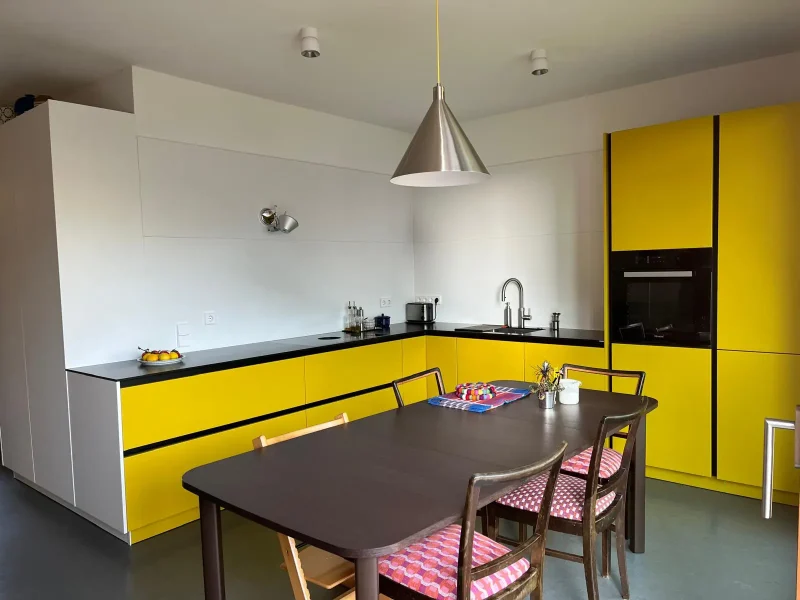 Moderne, zeitlos schöne Küche mit leuchtend gelben Schränken und schwarzen Arbeitsplatten, einem eingebauten Backofen, einem Esstisch mit unpassenden Stühlen und einer metallischen Pendelleuchte. Der Raum ist sauber, gut beleuchtet und wandelbar für jede Gelegenheit.