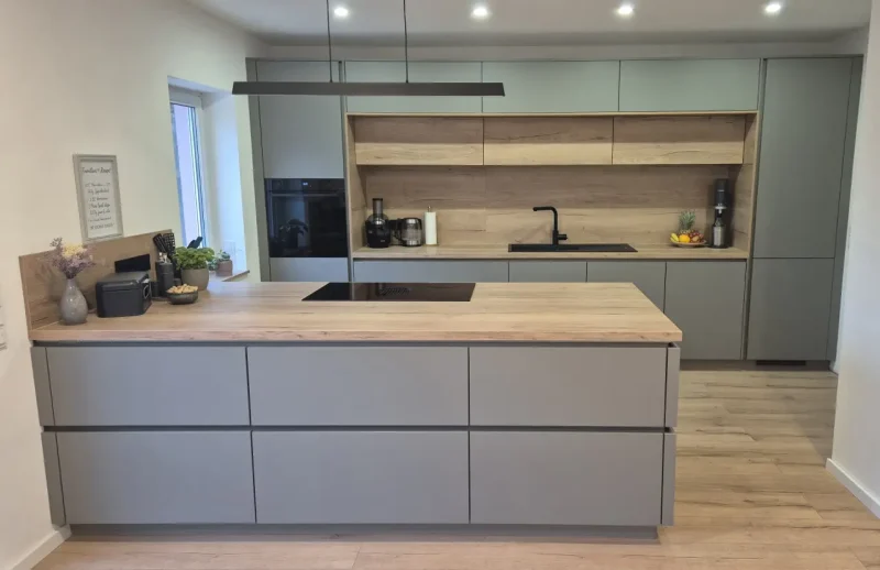 Moderne Küche mit hellgrauen Schränken, Arbeitsplatten aus Holz, einer schwarzen Spüle, Einbaugeräten und einer Insel mit Induktionskochfeld. Helle Küchen wie diese sind wandelbar und zeitlos schön - offen gestaltet mit Holzfußboden und offenen Regalen.