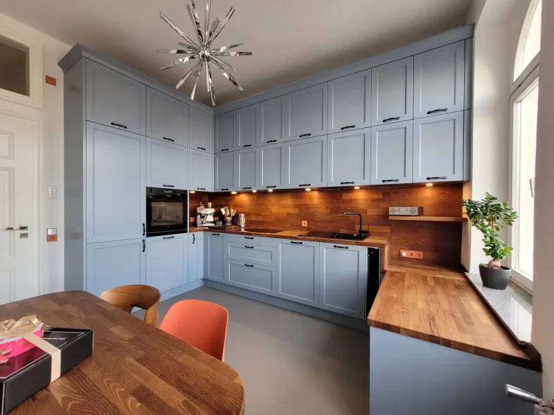 Moderne Küche mit hellblauen Schränken, Arbeitsplatten aus Holz, eingebautem Backofen und Unterschrankbeleuchtung. Diese helle Küche ist zeitlos schön, mit einem Holztisch im Vordergrund und einer Topfpflanze am Fenster unter einem modernen Kronleuchter.