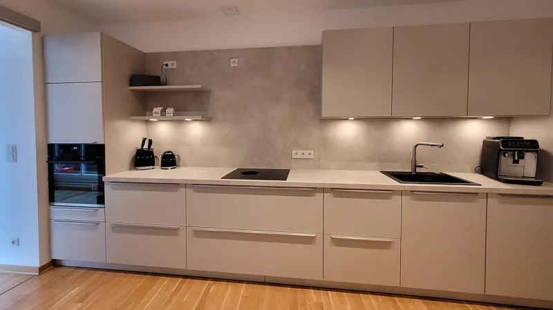 Moderne Küche mit beigen Schränken, Einbaubackofen, Induktionskochfeld, Unterbauleuchten und offenen Regalen auf einem hellen Holzboden. Die Helle Küchen Gestaltung ist zeitlos schön und verleiht dem minimalistischen Design eine wandelbare Eleganz.