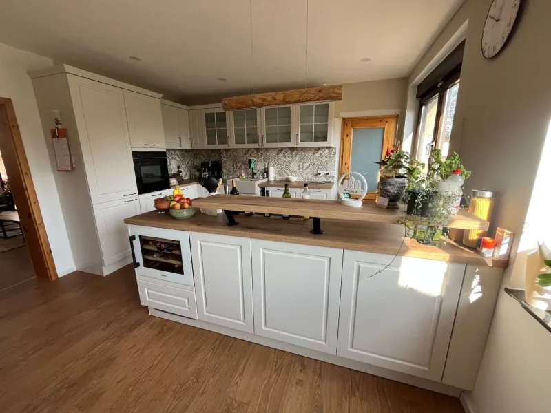 Helle, moderne Küche mit weißen Schränken, Holzarbeitsplatten und einer großen Insel. Dieser zeitlos schöne Raum ist mit Helle-Küchen-Details, Pflanzen und Obstschalen auf den Arbeitsflächen, einem eingebauten Ofen und Sonnenlicht, das durch ein Fenster einfällt, ausgestattet.