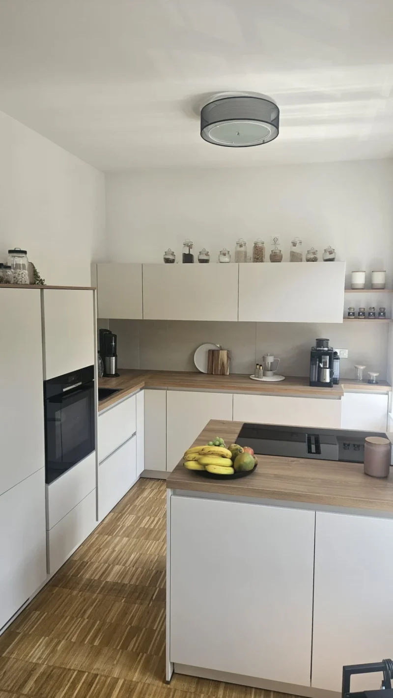 Moderne Küche mit weißen Schränken, Holzarbeitsplatten und eingebautem Backofen - ein Beispiel für helle Küchen. Auf der Theke steht eine Schale mit Obst, in den Regalen stehen Gläser und Dekoration. Geräte wie eine Kaffeemaschine stehen rechts. Heller Holzboden, natürliches Licht.