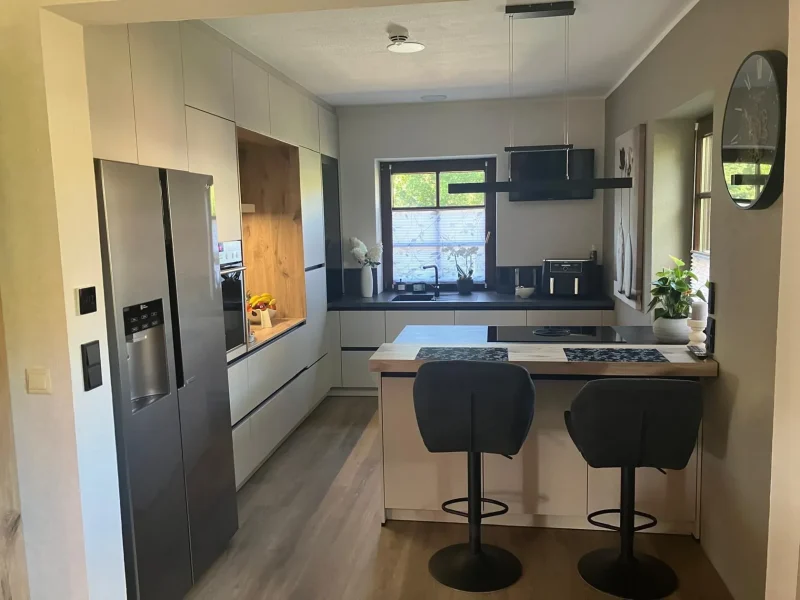 Moderne Küche mit hellen Küchen, hellen Schränken, einem Kühlschrank aus Edelstahl, Einbaubackofen, zwei schwarzen Barhockern an einer Insel, großen Fenstern, Topfpflanzen und Geräten auf der Theke. Eine zeitlos schöne Wanduhr hängt auf der rechten Seite.