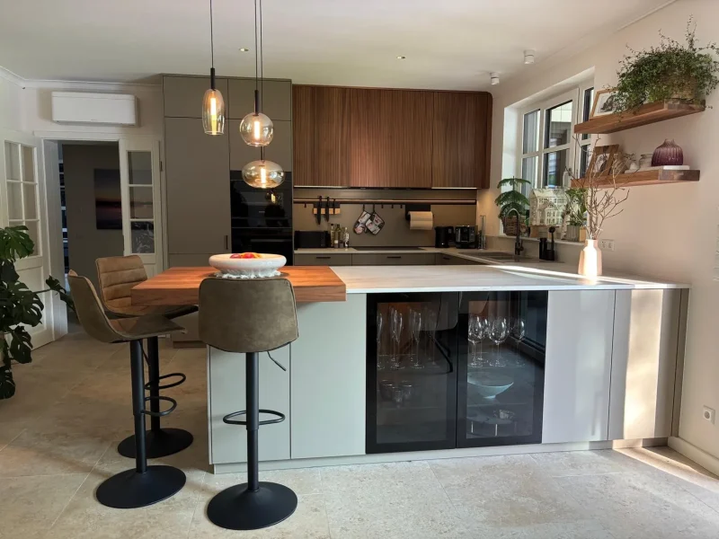 Moderne, wandelbare Küche mit großem Inselblock, zwei gepolsterten Barhockern, eingebauten Geräten und offenen Regalen mit Pflanzen und Dekor. Helle Küchen-Atmosphäre durch viel Tageslicht von den Fenstern über der Spüle.