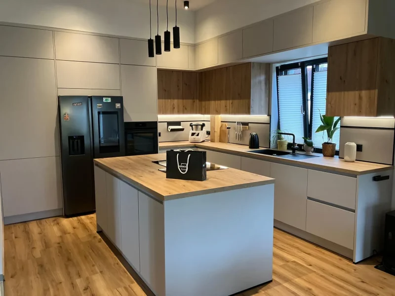 Moderne Küche mit hellem Holzfußboden, weißen Schränken, Holzarbeitsplatten, einer großen Kochinsel und einem Kühlschrank aus Edelstahl - diese Helle Küche ist zeitlos schön, mit schwarzen Pendelleuchten und Topfpflanzen, die den Fensterbereich erhellen.