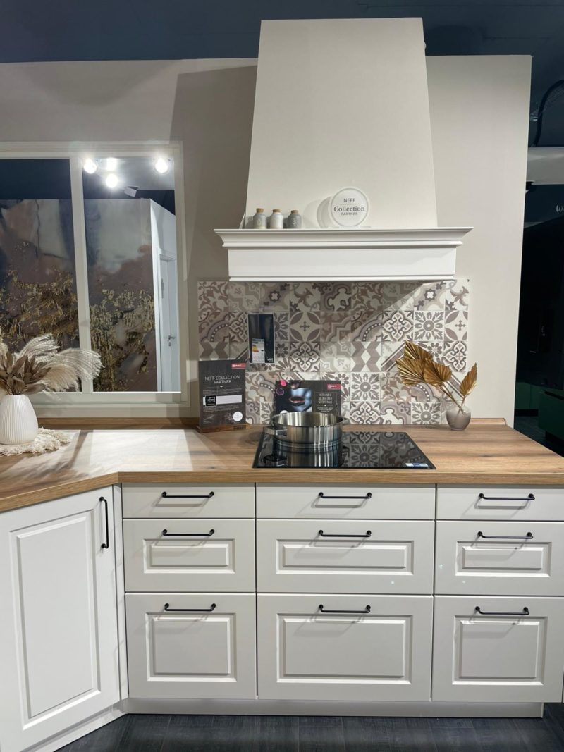 Moderne weiße Küche mit Holzarbeitsplatten, gemusterten Aufkantungen, einem eleganten Einbauherd Riva R 203, weißen Schränken mit schwarzen Griffen und dekorativen Gegenständen wie getrockneten Pflanzen und Xandora-Keramikgeschirr auf der Arbeitsplatte und im Regal.