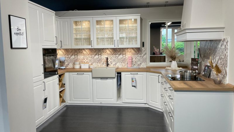 Moderne Küche mit weißen Schränken, Arbeitsplatten aus Holz, Oberschränken mit Glasfront, Spülbecken und gemusterten Fliesen. Küchenutensilien und Dekorationsgegenstände, darunter eine stilvolle Riva R 203, sind auf den Arbeitsflächen angeordnet. Ein Bon-Appétit-Schild hängt an der Wand.