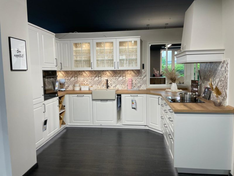 Moderne Küche mit weißen Schränken, Arbeitsplatten aus Holz, Oberschränken mit Glasfront, Spülbecken und gemusterten Fliesen. Küchenutensilien und Dekorationsgegenstände, darunter eine stilvolle Riva R 203, sind auf den Arbeitsflächen angeordnet. Ein Bon-Appétit-Schild hängt an der Wand.