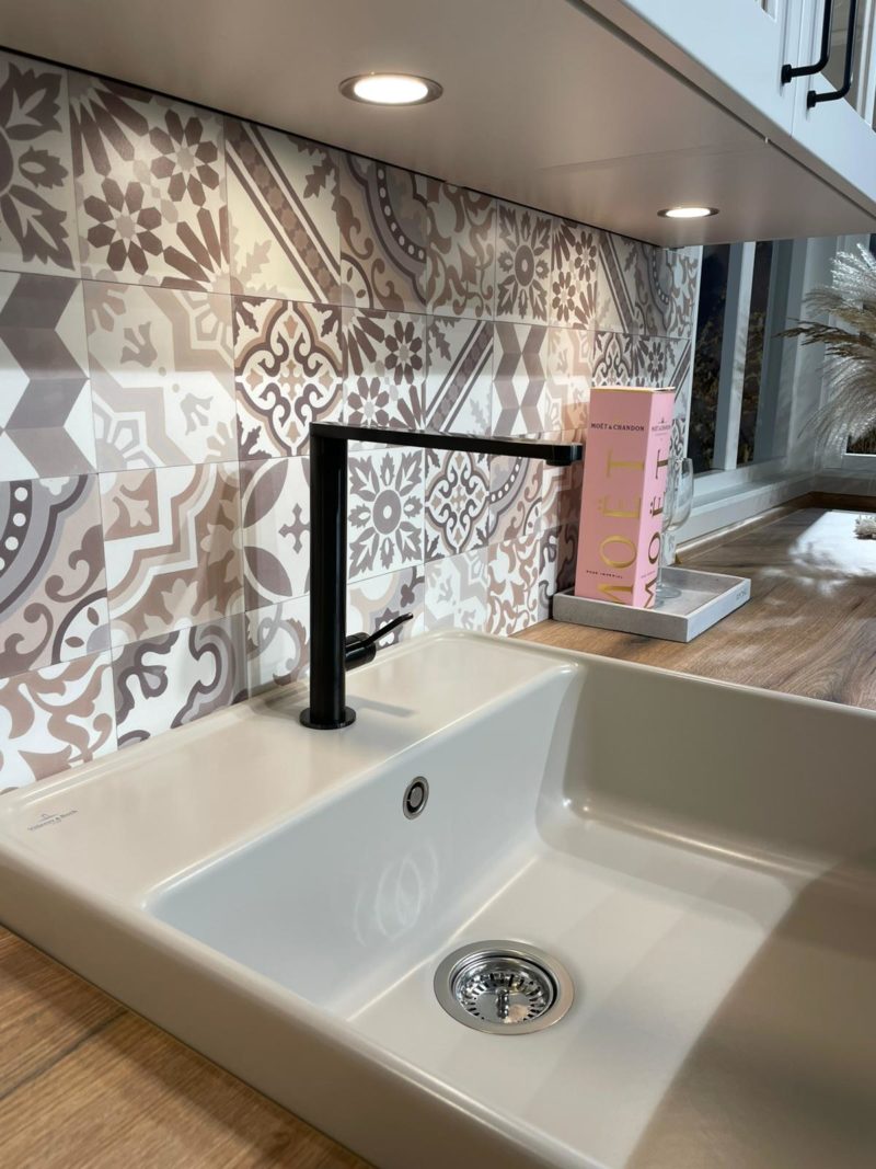 Eine moderne Küchenspüle mit einer mattschwarzen Riva R 203-Armatur sitzt unter einer gemusterten Fliesenrückwand und Unterbauleuchten. Auf der hölzernen Arbeitsplatte stehen eine pinkfarbene Moët-Dose und Pampasgras-Dekor.