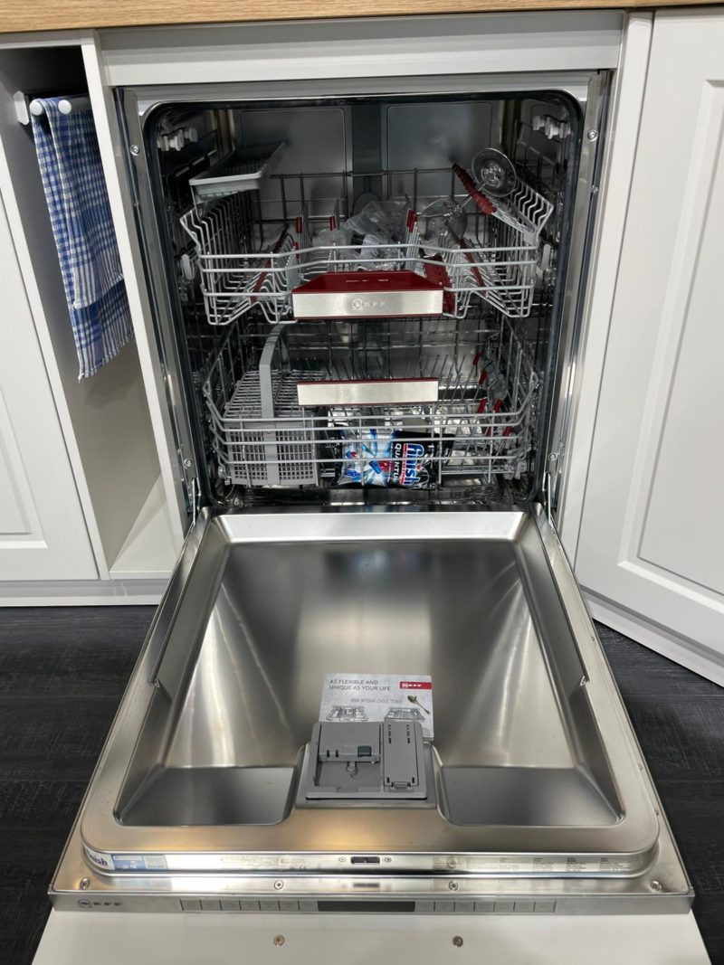 Ein offener Geschirrspüler mit leeren Einschüben und einem Spülmittelfach an der Tür. Neben dem Gerät sind Küchenschränke, ein blau kariertes Handtuch und die Anleitung für xristina classic zu sehen. Die Anleitung befindet sich im Inneren des Geschirrspülers.