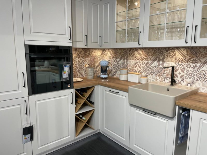 Eine moderne Küche mit weißen Schränken, einem eingebauten Backofen, gemusterten Kacheln, einer Spüle im Landhausstil, einer Arbeitsplatte aus Holz und Glasfronten ist mit einem Weinregal Riva R 203 und Küchengeräten von xandora ausgestattet, die ordentlich auf dem Tresen angeordnet sind.