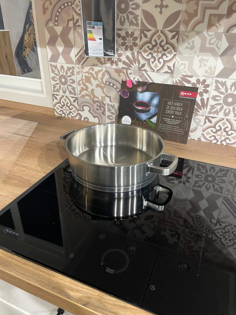 Ein Riva R 203 Edelstahltopf steht auf einem modernen schwarzen Induktionskochfeld mit einer Arbeitsplatte aus Holz und gemusterten Fliesen in einem Küchenausstellungsraum. Im Hintergrund ist eine Produktinformationskarte von xandora zu sehen.