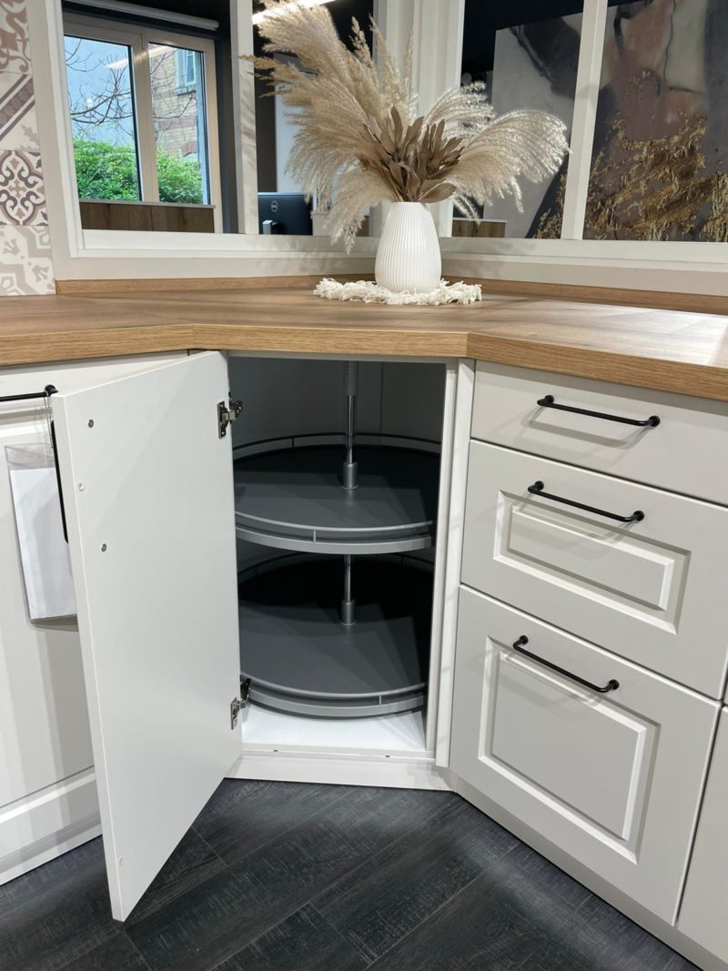 Ein weißer Xandora-Küchen-Eckschrank mit offener Tür gibt den Blick frei auf ein zweistöckiges, drehbares Regal, das neben Schubladen steht. Auf einer hölzernen Arbeitsplatte steht eine weiße Vase mit getrocknetem Pampasgras, die das moderne Design von Riva R 203 unterstreicht.