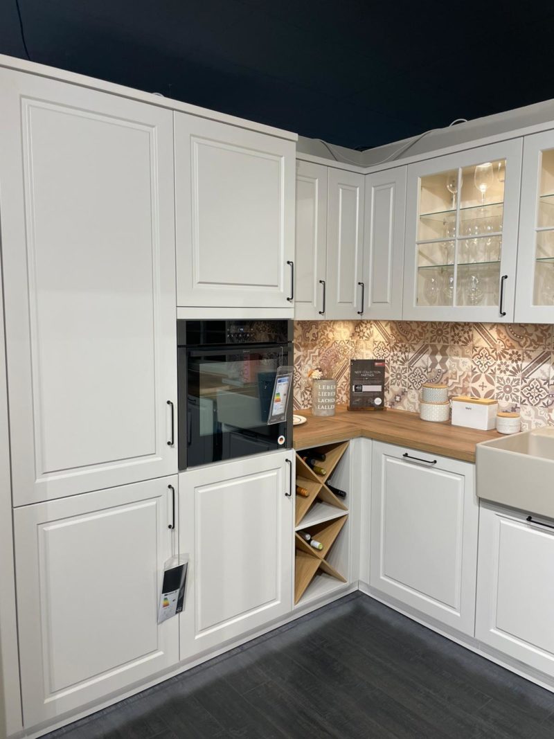 Eine moderne Küchenecke mit weißen Riva R 203-Schränken, einer gemusterten Aufkantung, einem eingebauten Backofen, einem Oberschrank mit Glasfront, einer Arbeitsplatte aus Holz, einem Xandora-Weinregal und dekorativen Gläsern auf der Theke.