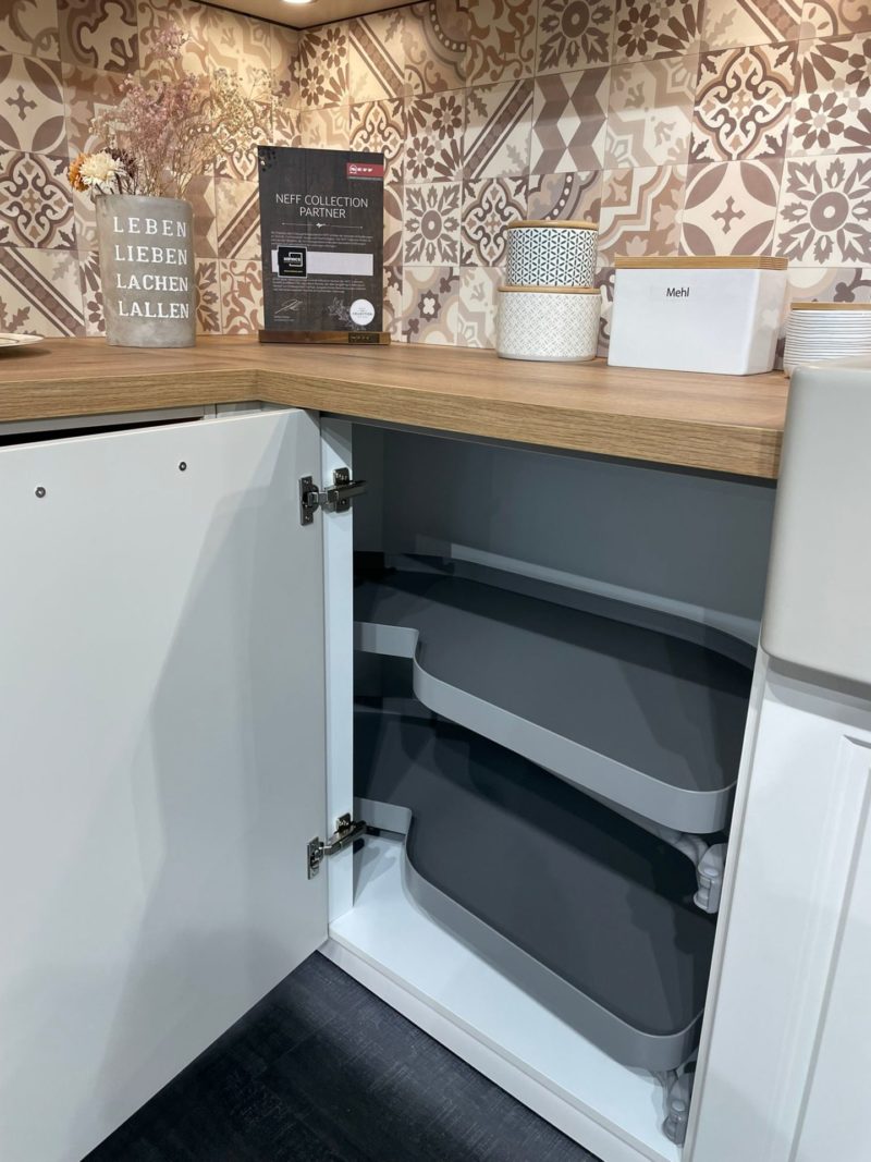 Ein moderner Kücheneckschrank Riva R 203 ist geöffnet und gibt den Blick auf zwei graue, ausziehbare Regale frei. Auf der hölzernen Arbeitsplatte darüber befinden sich dekorative Fliesen, eine Vase mit Trockenblumen, ein weißer Kanister und ein Serviettenhalter mit der Aufschrift Mein.