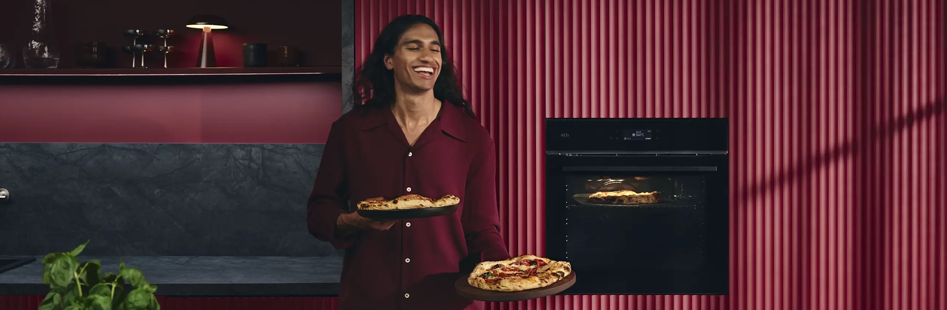 Eine Person in einem roten Hemd lächelt, während sie zwei Pizzen in einer modernen roten Küche hält. Eine weitere Pizza backt im Ofen hinter ihnen. Das Ambiente ist geprägt von dunklen Arbeitsflächen und rot gestreiften Wänden.