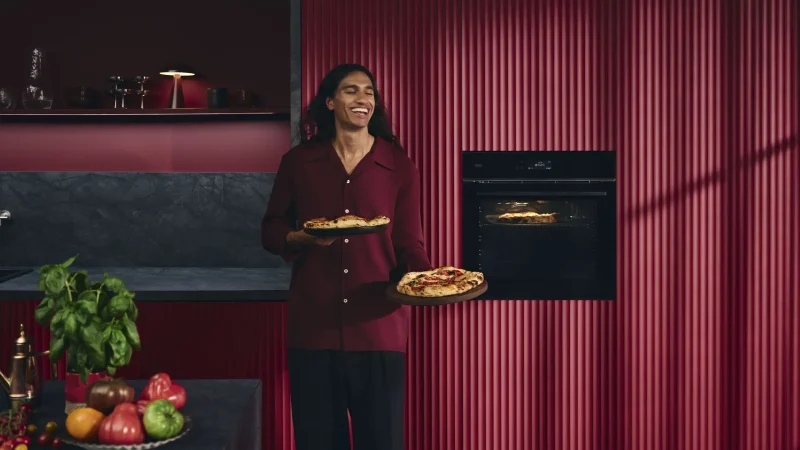 Eine Person steht lächelnd in einer modernen Küche mit dunklen Arbeitsflächen und roten Wänden und hält zwei Pizzen in der Hand. Eine weitere Pizza backt im Backofen hinter ihnen. Frisches Gemüse und Kräuter ergänzen die Genussmomente auf der Küchentheke.