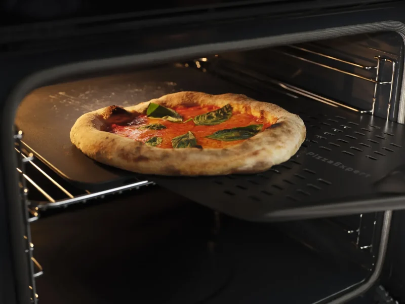 Eine Pizza Margherita mit Tomatensauce und Basilikumblättern wird auf einem schwarzen Pizzaschieber aus dem Backofen geholt, ihre dicke, goldbraune Kruste verspricht echte xanocs Genussmomente.