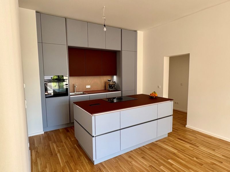 Moderne, minimalistische Küche mit deckenhohen, grauen Schränken kombiniert mit einer braunen Kochinsel samt Induktionskochfeld, Einbaubackofen, Kaffeemaschine und Obstschale. Holzfußboden und neutrale Wände runden den Look ab.