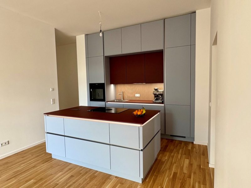 Eine moderne Küche mit deckenhoch kombinierten, lichtgrauen Schränken, einer braunen Kochinsel mit Induktionskochfeld, eingebautem Ofen, Spüle und Obstschale. Der Boden ist helles Holz, eine einzelne Glühbirne hängt von der Decke.