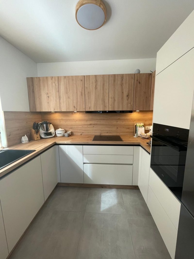 Moderne Wohnküche mit weißen Schränken, Holzoberschränken und einer Rückwand aus Holz. Die Stauraumlösung sorgt für Ordnung, während Induktionskochfeld, Einbauherd und schwarze Spüle Funktionalität garantieren. Graue Fliesen runden das Design ab.