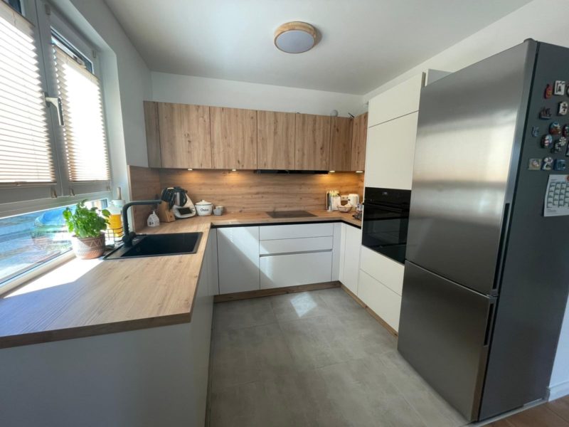 Moderne Wohnküche mit Holz- und weißen Schränken, schwarzer Spüle, Stauraumlösung, Einbaubackofen, Edelstahl-Kühlschrank mit Magneten, Küchenutensilien auf der Arbeitsplatte und Fenstern mit Jalousien. Eine Topfpflanze ziert die Fensterbank.