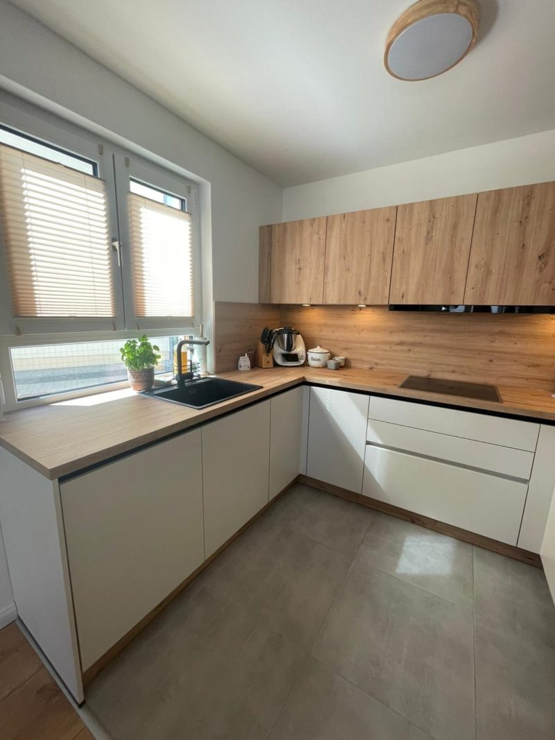 Moderne Wohnküche mit Stauraumlösung: Lichtdurchflutete Küche mit hellen Holz- und weißen Unterschränken, schwarzem Spülbecken, Induktionskochfeld sowie kleinen Geräten auf der Arbeitsplatte. Am Spülbecken steht eine Pflanze.