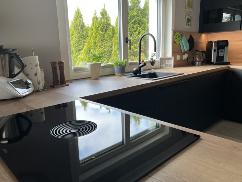 Moderne Küche mit Arbeitsplatte aus Holz, elegante schwarze Schränke, Induktionskochfeld, Spüle mit Armatur, Kaffeemaschine, Zimmerpflanzen am großen Fenster und viel Tageslicht – perfekt für Ihr neues Zuhause.