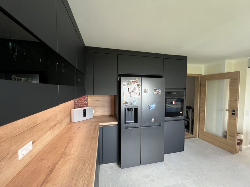 Eine moderne Küche mit mattschwarzen Schränken, Arbeitsplatten und Fliesenspiegel aus Holz, einem Kühlschrank mit Magneten und Fotos, einem Backofen und einer hellen Tür mit Glaseinsätzen – schwarze Eleganz für dein neues Zuhause. Der Raum ist aufgeräumt und übersichtlich.