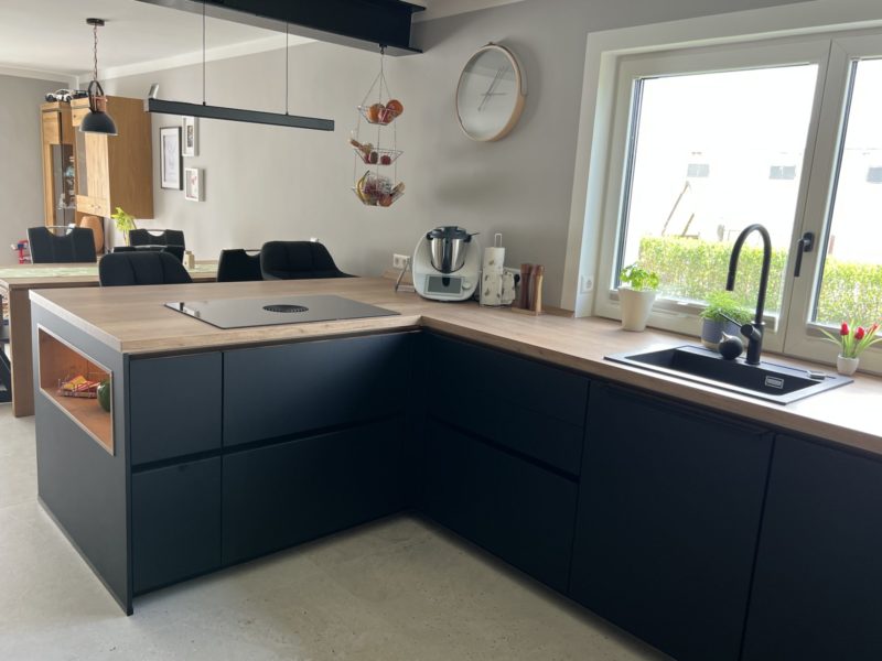 Moderne Küche mit schwarzer Eleganz: Schwarze Schränke, Arbeitsplatten aus Holz, Induktionskochfeld und Spüle am großen Fenster. Eine Uhr, Pflanzen und Küchengeräte sorgen für wohnliches Flair im neuen Zuhause; im Hintergrund der Essbereich.
