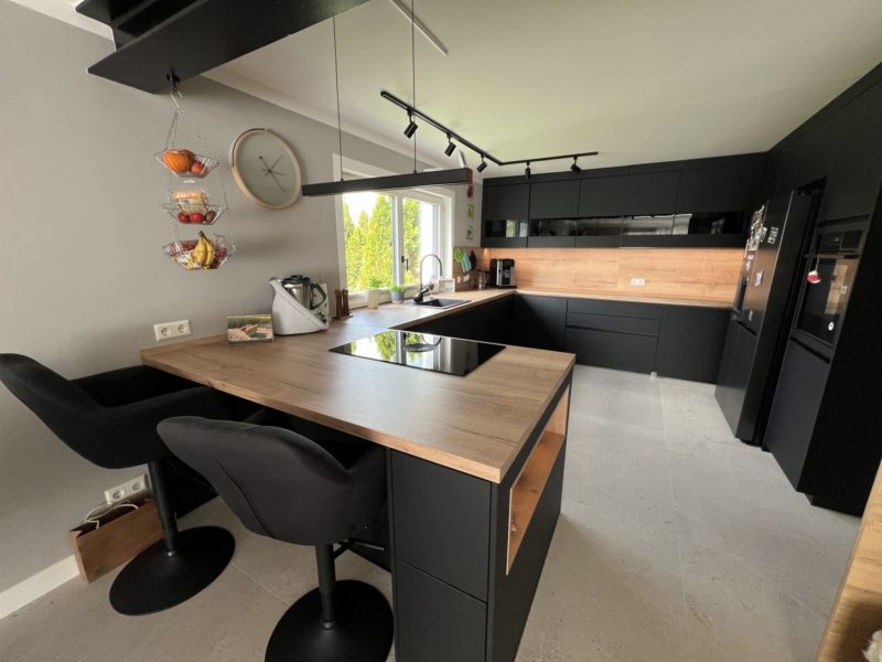 Die moderne Küche mit schwarzen Schränken, Arbeitsplatten aus Holz, Einbaugeräten und einer Kochinsel mit zwei schwarzen Stühlen strahlt schwarze Eleganz aus. Ein hängender Obstkorb und ein großes Fenster schaffen einen hellen, aufgeräumten Raum für Ihr neues Zuhause.