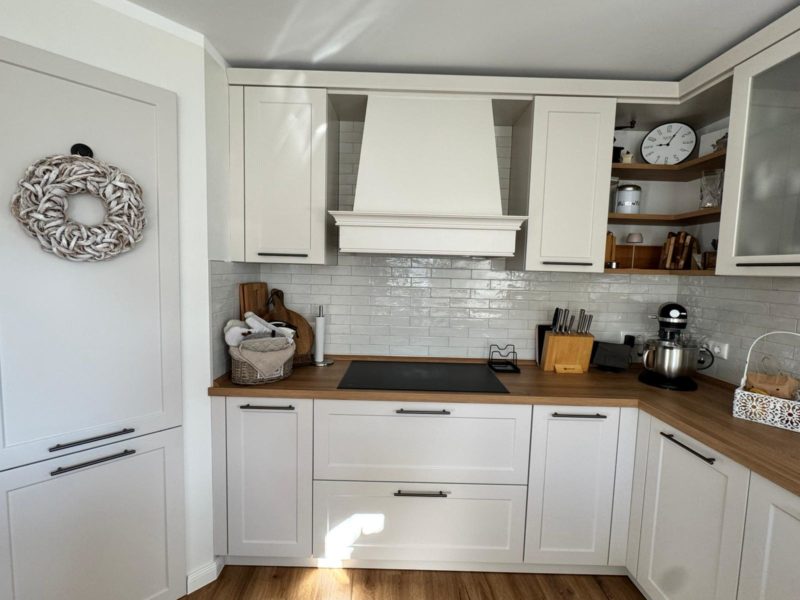 Helle, moderne Landhausküche mit weißen Schränken, Arbeitsplatten aus Holz und einer weißen Fliesenrückwand. Dank reichlich Stauraum lassen sich Küchengeräte, Küchenutensilien, eine Küchenmaschine sowie Dekoartikel wie eine Wanduhr und ein Kranz ordentlich unterbringen.