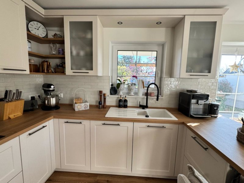 Eine moderne Landhausküche mit weißen Schränken, Arbeitsplatten aus Holz und einer weißen Fliesenrückwand. Ein Fenster über der Spüle bietet Blick auf den Garten, während dank cleverer Stauraumlösungen Küchengeräte, Küchenutensilien und die Kaffeemaschine ordentlich verstaut sind.