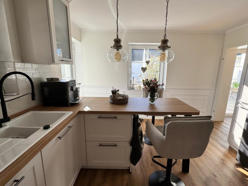 Helle, moderne Landhausküche mit weißen Schränken, Arbeitsplatten aus Holz und einer kleinen Kücheninsel mit einem beigen Stuhl. Darüber hängen zwei kugelförmige Pendelleuchten, und auf der Kücheninsel steht eine Vase mit Blumen neben einem Fenster, durch das Sonnenlicht hereinscheint. Zusätzlicher Stauraum ist vorhanden.