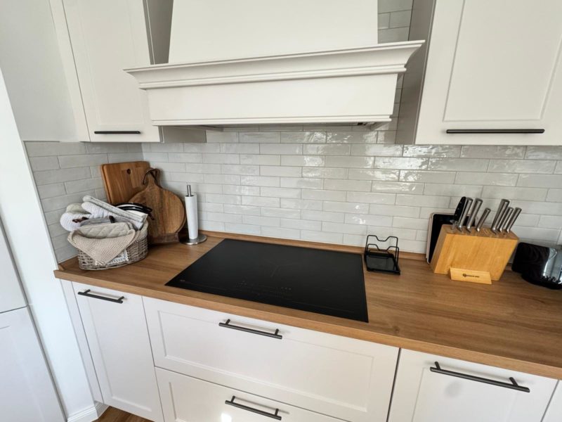 Moderne Küche mit weißen Schränken, einer Arbeitsplatte aus Holz, einem Induktionskochfeld, einem Messerblock, einem Küchenrollenhalter, einem Besteckkorb, Schneidebrettern und einer weißen Fliesenrückwand im Subway-Stil – perfekter Stauraum für eine stilvolle Landhausküche.