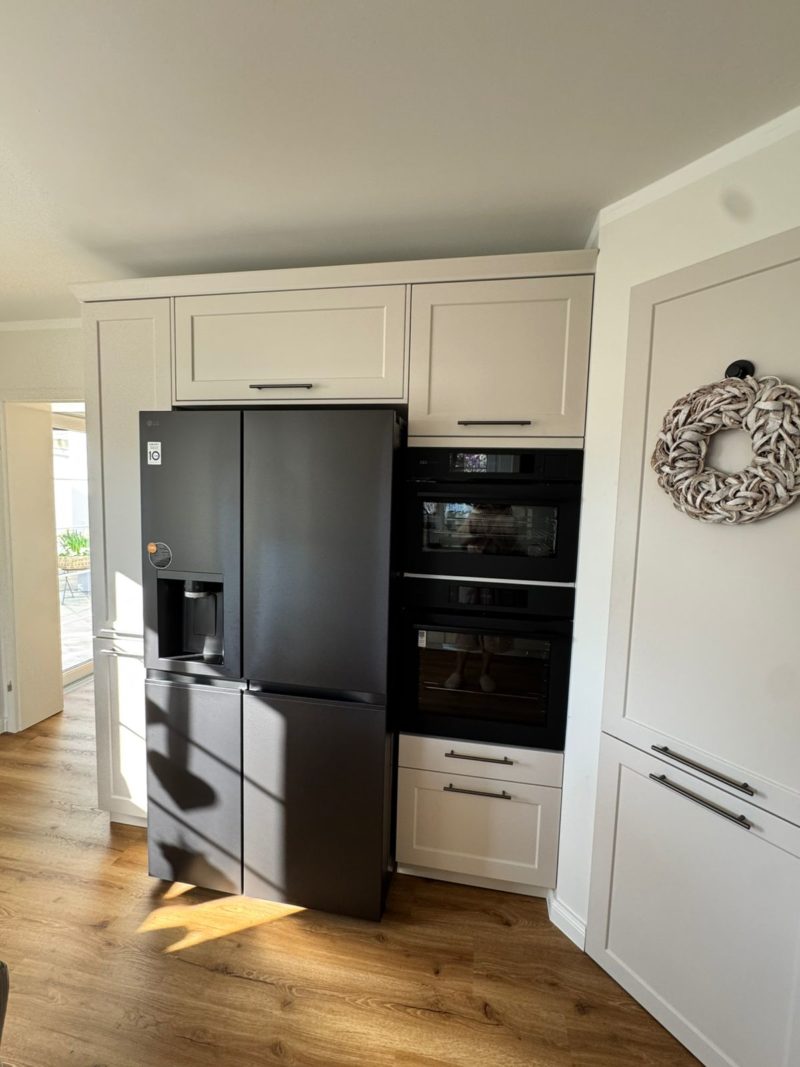 Eine moderne Landhausküche mit hellen Schränken, einem großen schwarzen zweitürigen Kühlschrank mit Wasserspender, Einbaubacköfen und einem dekorativen Kranz an einer weißen Wand. Sonnenlicht fällt durch eine nahegelegene Tür herein und bringt den großzügigen Stauraum zur Geltung.