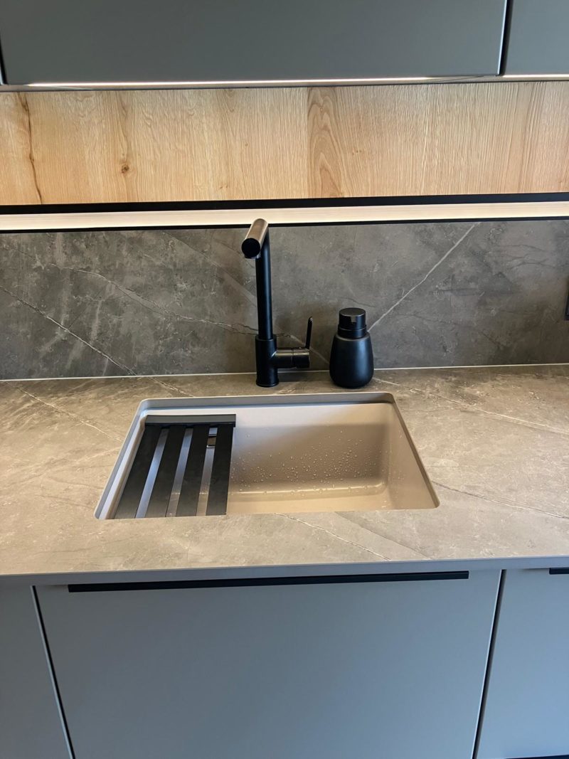 Eine moderne Küchenspüle mit einer eleganten schwarzen Armatur und einem Seifenspender ist in eine graue Stein-Arbeitsplatte eingelassen und fügt sich nahtlos in die offene Wohnküche ein. Graue Schränke und ein Regalbock aus Holz darüber unterstreichen den Wohlfühlcharakter des Raums.