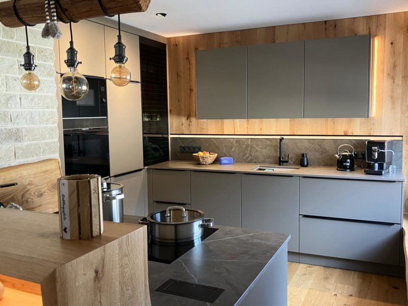 Moderne offene Wohnküche mit mattgrauen Schränken, Holzakzenten, Einbaugeräten und einer Marmorarbeitsplatte. Pendelleuchten über der Kücheninsel und ein durchdachter Grundriss sorgen für ein stilvolles, aufgeräumtes Wohlfühlambiente.