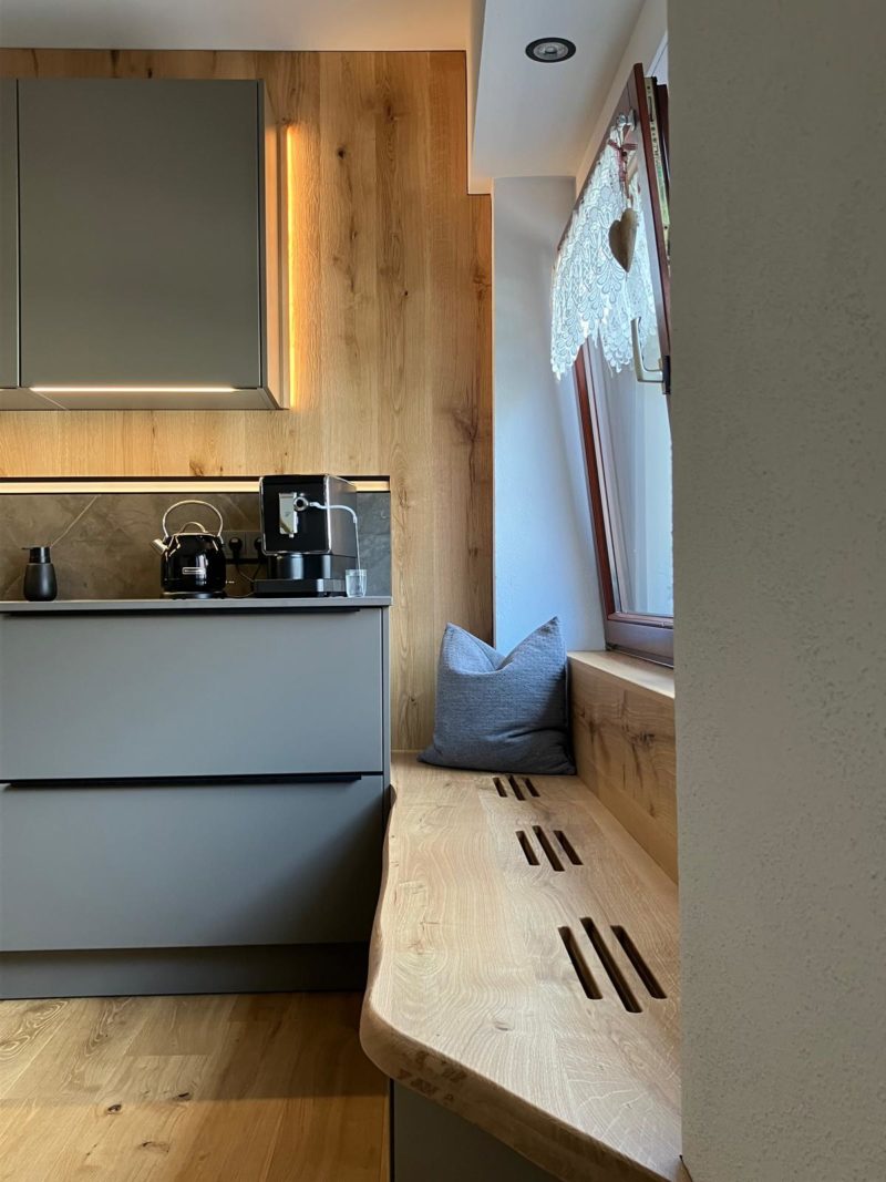 Moderne Küche mit grauen Schränken und Holzoberflächen, als offene Wohnküche gestaltet. Wasserkocher und Kaffeemaschine stehen bereit. Eine eingebaute Holzbank mit Kissen unter dem Fenster mit Spitzenvorhang und Herzdekoration unterstreicht den Wohlfühlcharakter.