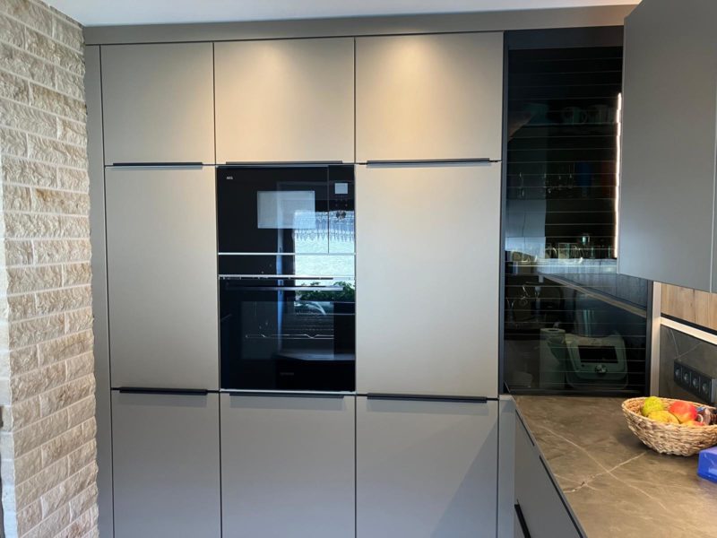 Eine elegante, moderne Küche mit mattgrauen Schränken, einem eingebauten schwarzen Backofen und einer hellen Steinwand – diese offene Wohnküche besticht durch eine graue Arbeitsplatte mit Obst- und Glasregalen auf der rechten Seite, die für echten Wohlfühlcharakter sorgen.