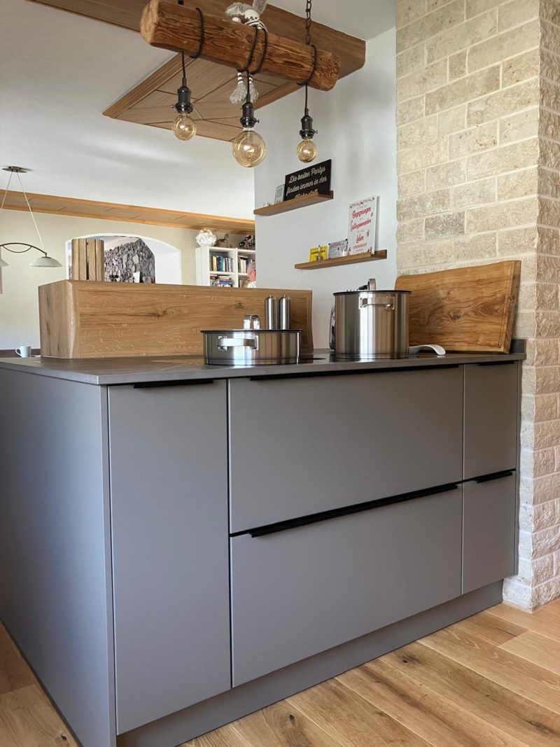 Moderne offene Wohnküche mit mattgrauen Schränken, Holzakzenten und einer hängenden Holzlampe mit sichtbaren Glühbirnen. Küche mit Wohlfühlcharakter: Topf, Gewürzregal und Schneidebretter auf der Kücheninsel, helle Holzböden und eine freiliegende Ziegelwand.