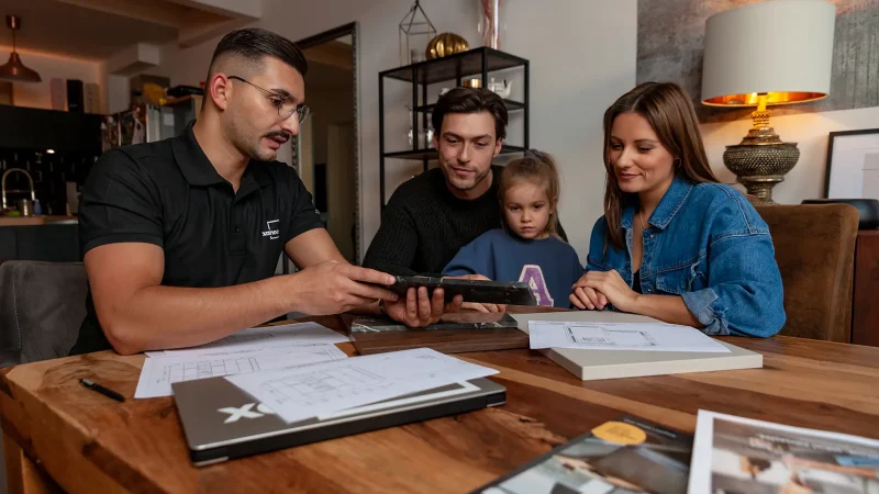 Ein Mann in einem schwarzen Hemd bietet einem Paar und ihrer kleinen Tochter an einem Holztisch, umgeben von Papieren, einem Laptop und Broschüren, in einer gemütlichen, modernen Wohnumgebung eine gute Küchenberatung an.