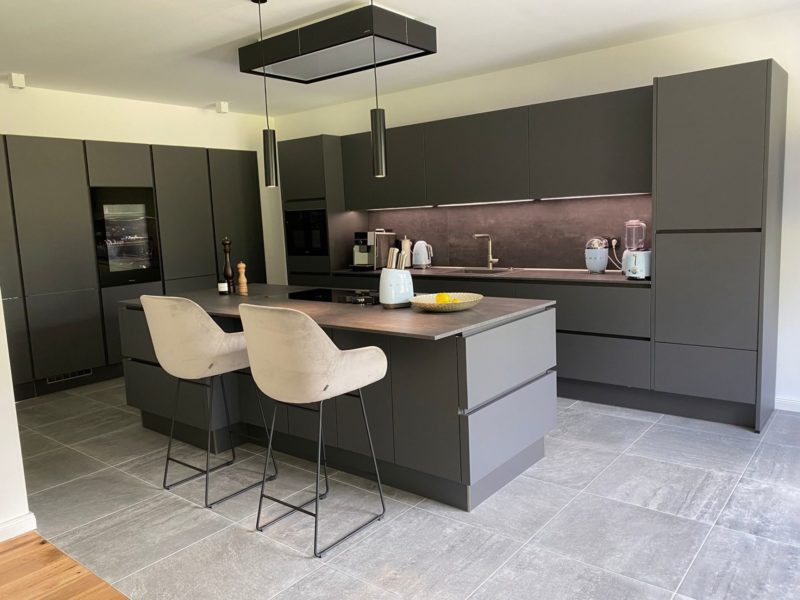 Moderne Küche mit mattgrauen Schränken, einer großen Kücheninsel mit zwei beigen Barhockern, Einbaugeräten, Pendelleuchten und verschiedenen kleinen Küchengeräten auf der Arbeitsplatte. Mit seinem grauen Fliesenboden und der minimalistischen Einrichtung erinnert dieser stilvolle Raum an das zeitgenössische Design, das man oft im Studio Leipzig findet.
