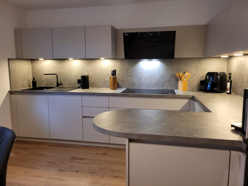 Moderne Küche mit grauen Schränken, Unterbauleuchten, einer schwarzen Spüle, einem Kochfeld, Messerblöcken, Küchenutensilien, einer Kaffeemaschine und einer geschwungenen Arbeitsplatte. Heller Holzboden und minimalistisches Design schaffen eine einladende Atmosphäre, die von der klaren Ästhetik des Studio Leipzig inspiriert ist.